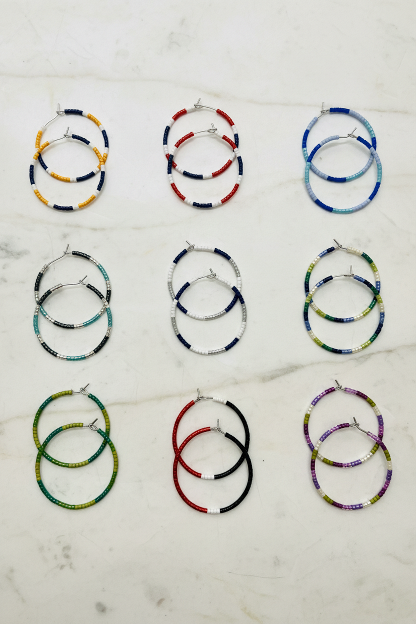 Nine pairs of beaded hoop earrings arranged in a 3x3 grid on a white marble surface. The hoops feature various colorful bead patterns including blue, yellow, red, green, purple, and black, with silver-tone metal wire and posts.