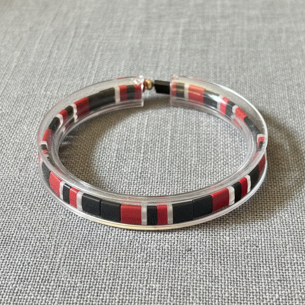 A clear acrylic bangle bracelet featuring a striped inlay of red, black, and white square beads, resting on a dark wooden surface.