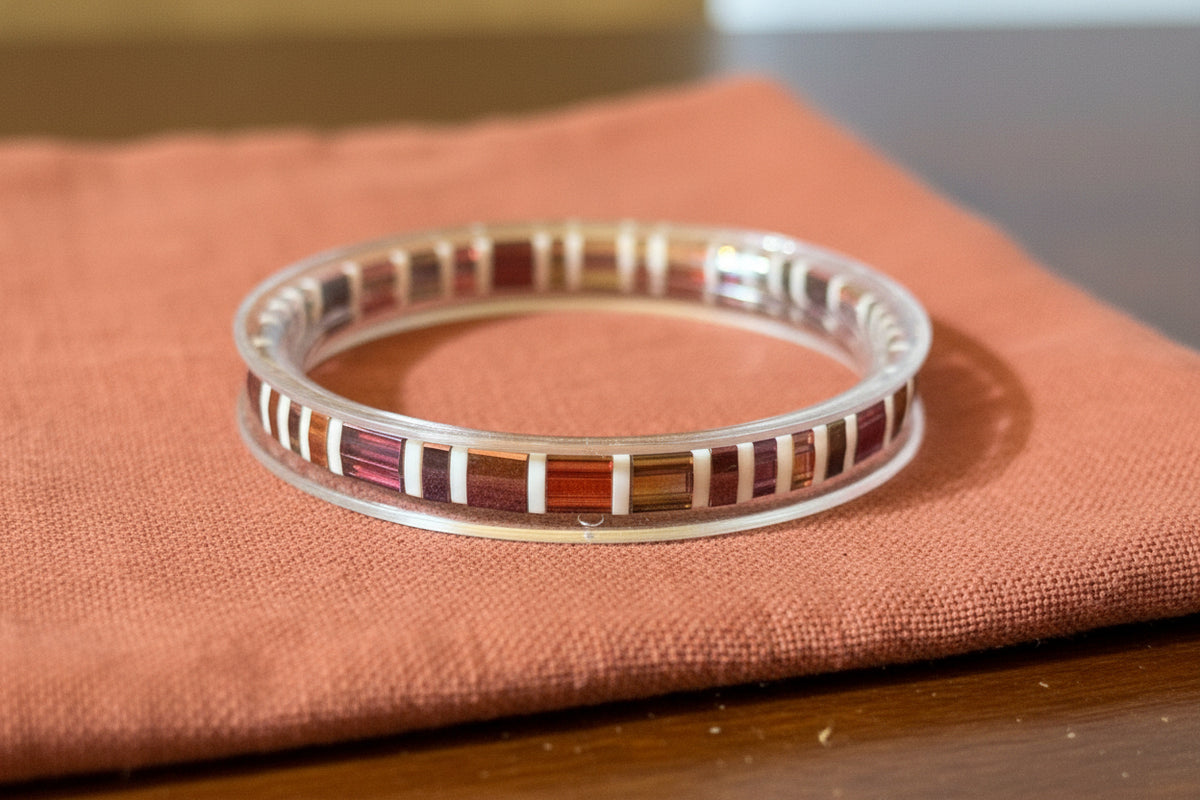 A clear acrylic cuff bracelet with alternating inlaid stripes of reddish-brown wood and white material, resting on a dark wooden table.