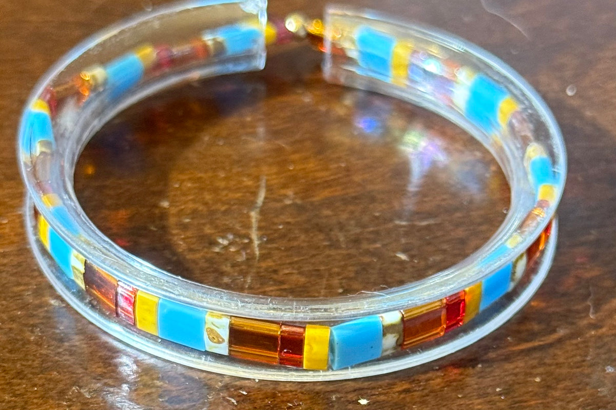 A clear acrylic bangle bracelet featuring rectangular turquoise blue, yellow, and amber glass inlays, resting on a dark wooden surface.