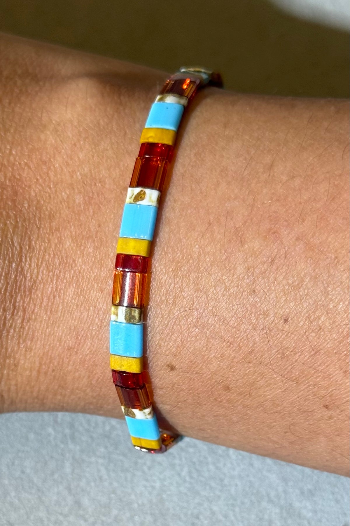 A close-up of a beaded stretch bracelet worn on a person's wrist. The bracelet features alternating translucent amber-orange and bright light blue rectangular beads, separated by thin yellow stripes.