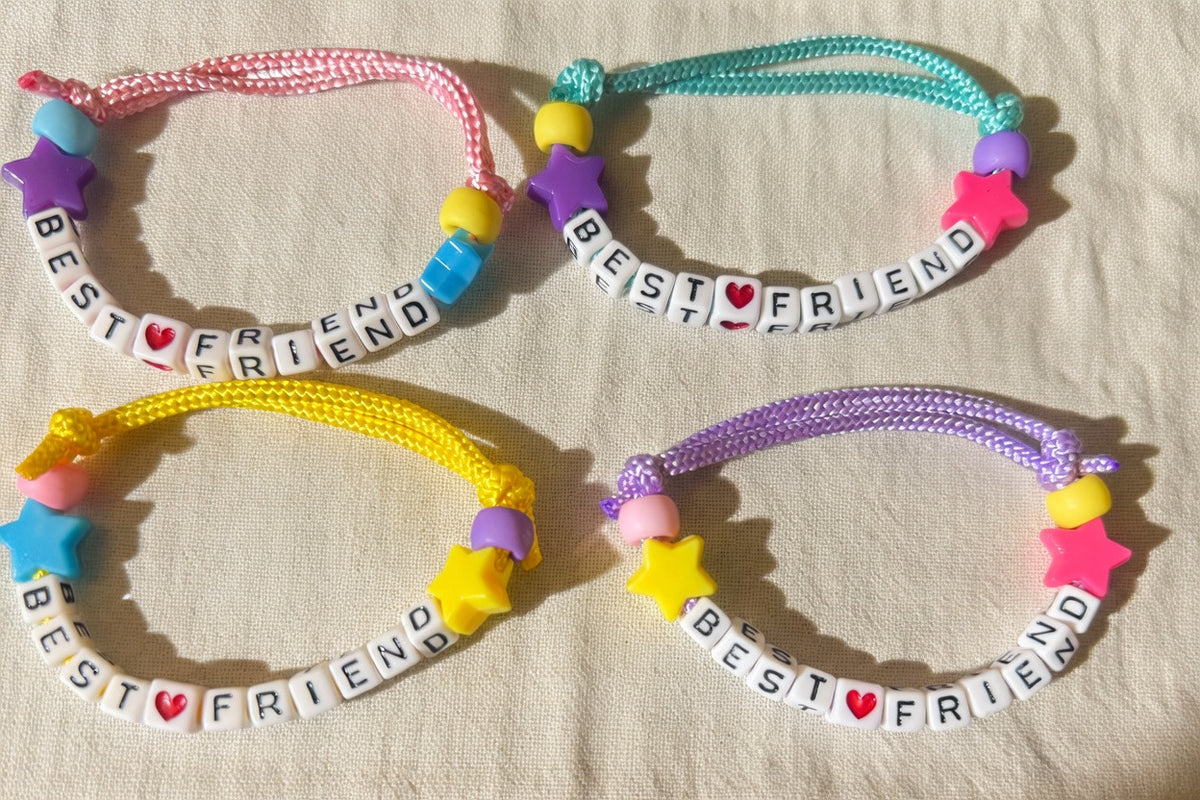 Four handmade friendship bracelets arranged in a 2x2 grid on a beige fabric background. Each bracelet features a braided cord, a purple star charm, a pink and white heart charm, a yellow star charm, and white letter beads spelling 'BEST FRIEND' with a small red heart.