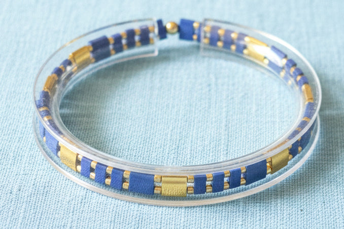 A clear acrylic cuff bracelet with alternating blue and gold rectangular beads, resting on a dark wooden surface.