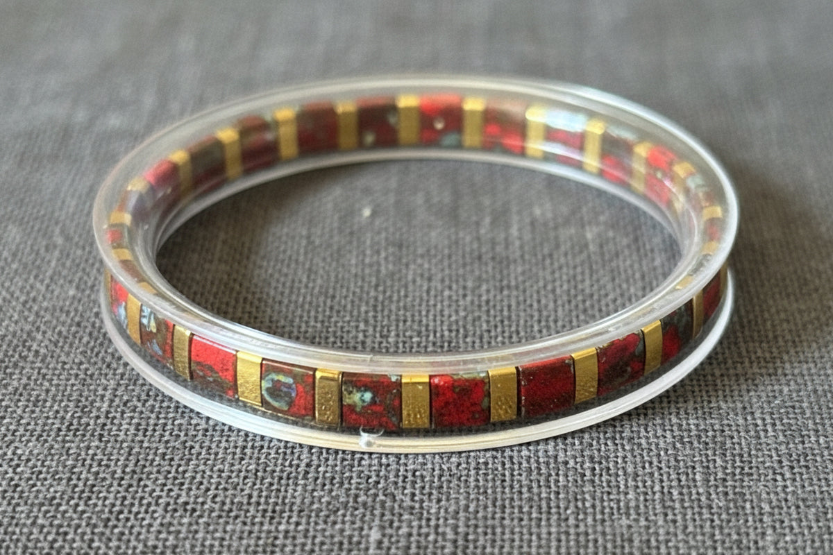 A clear acrylic bangle bracelet with a mosaic inlay of red, black, and gold square tiles, resting on a reflective wooden surface.