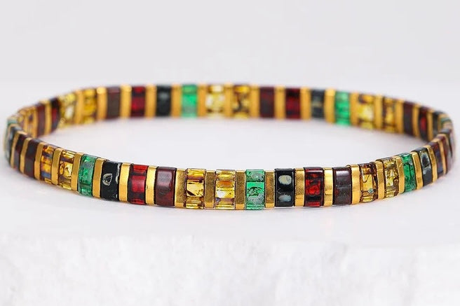 A gold-plated bangle bracelet featuring alternating segments of green emerald-cut stones, red baguette-cut stones, and black rectangular stones, displayed on a white textured surface.