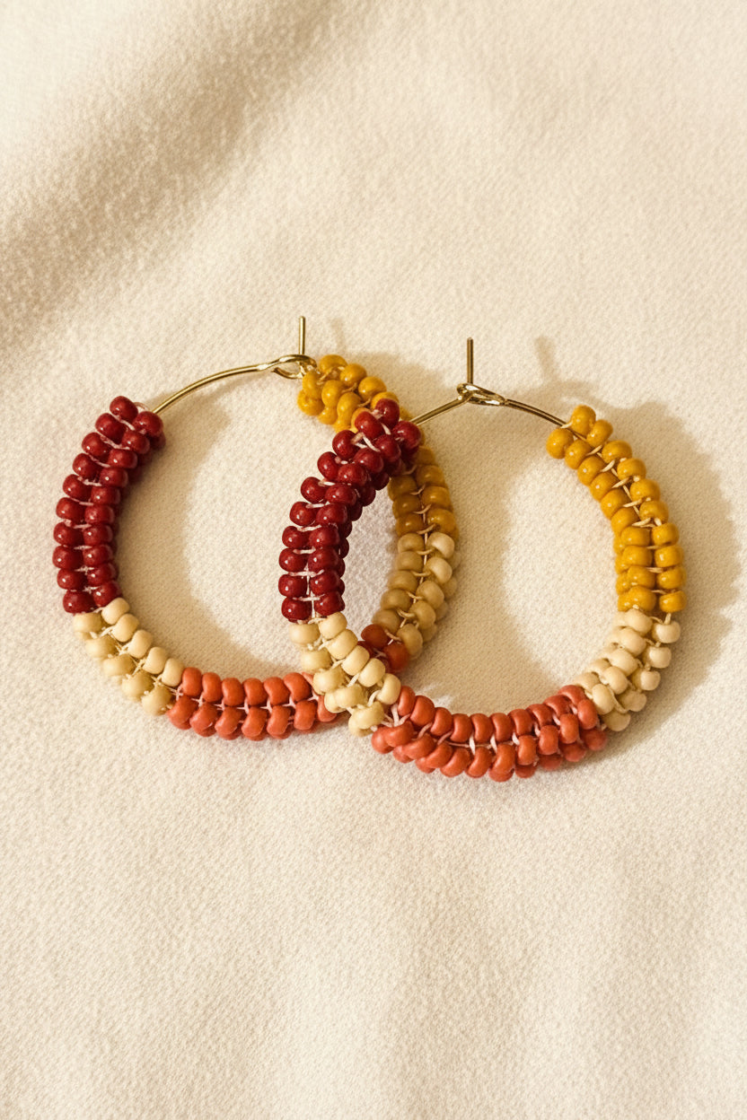 Three beaded hoop earrings with a warm autumn color palette, featuring red, orange, beige, and mustard yellow beads on a soft cream fabric background.