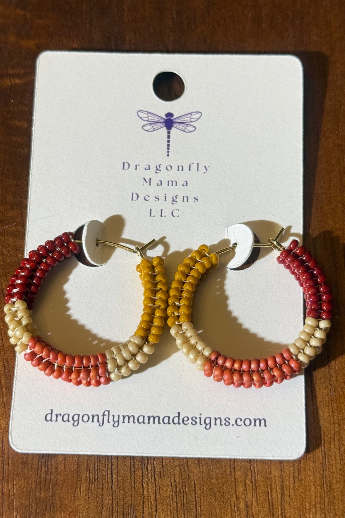 A pair of beaded hoop earrings displayed on a white brand card over a wooden surface. The earrings have three rows of beads in red, orange, and cream, with gold-tone posts and white backings.